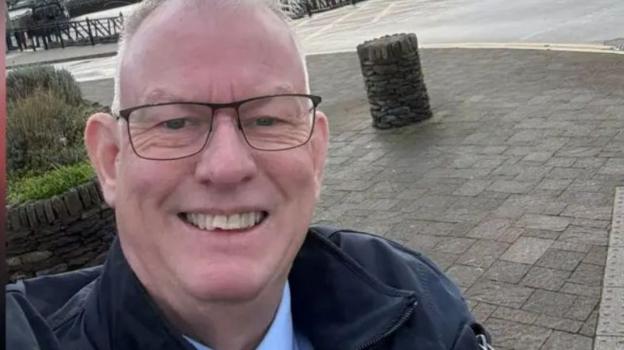 A selfie of Elfyn Thomas smiling as he stands on a paved area close to a dockside. He's wearing glasses, a blue shirt and a jacket.
