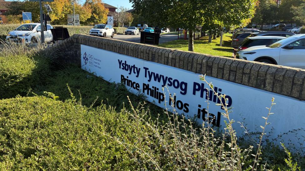 A hospital sign that says 'Ysbyty Tywysog Philip - Prince Philip Hospital'. The 'p' is missing in the word hospital on the sign. It is surrounded by greenery and cars.