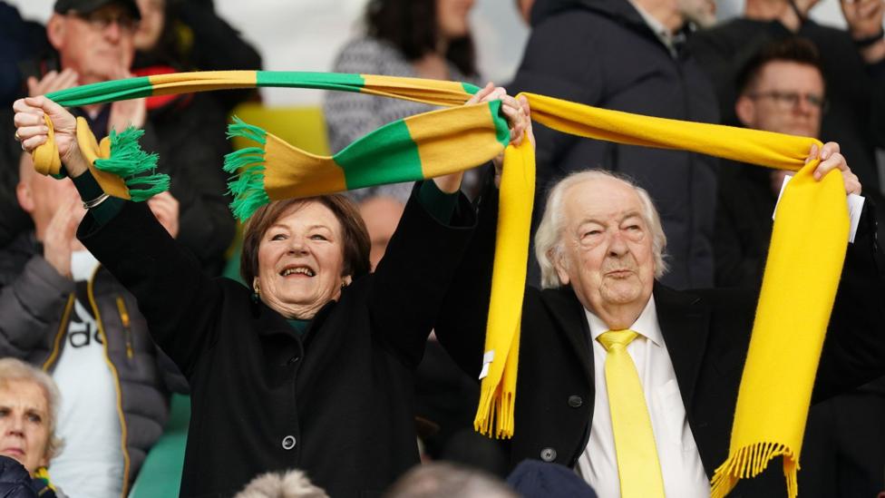 Delia Smith and Michael Wynn Jones holding Norwich City scarves aloft at a game. They are both dressed smartly and smiling.