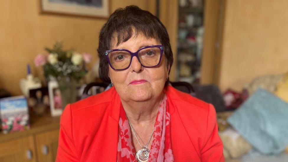 Teresa is short dark hair, glasses, a red jacket over a red floral pattern top. She is sitting in a room in a house.