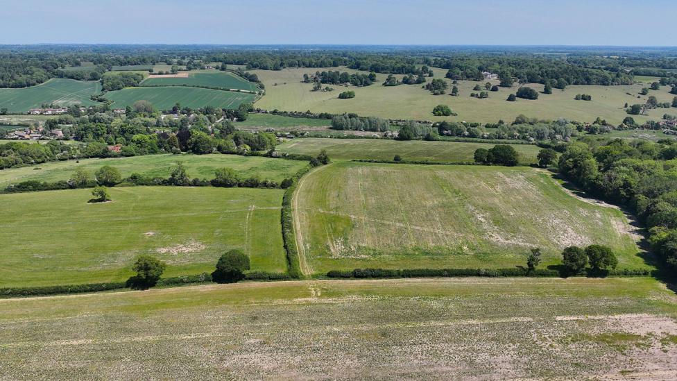 A large arable farm area, with trees in it, carved into different sections. There are houses to the left.