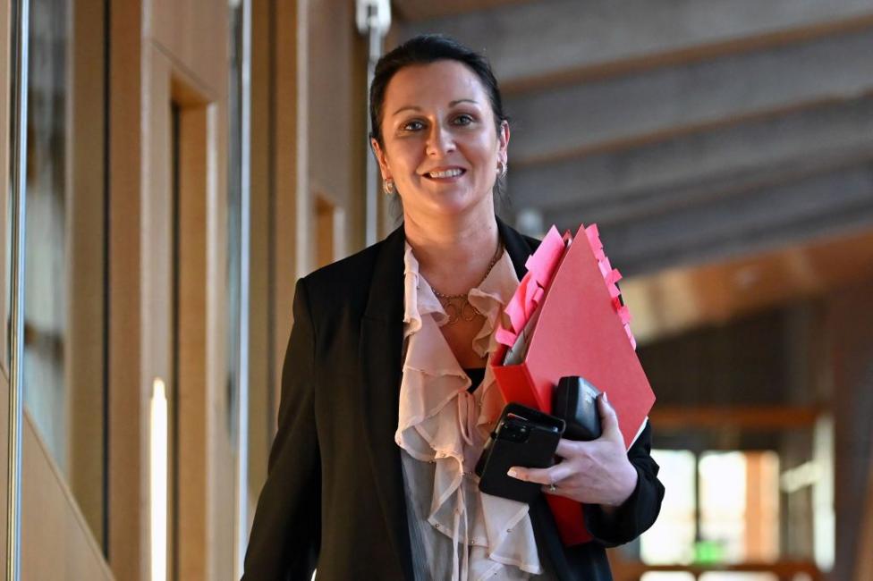 A woman with dark hair, pulled back smiles. She is walking while holding a folder of papers, a phone and a glasses case.