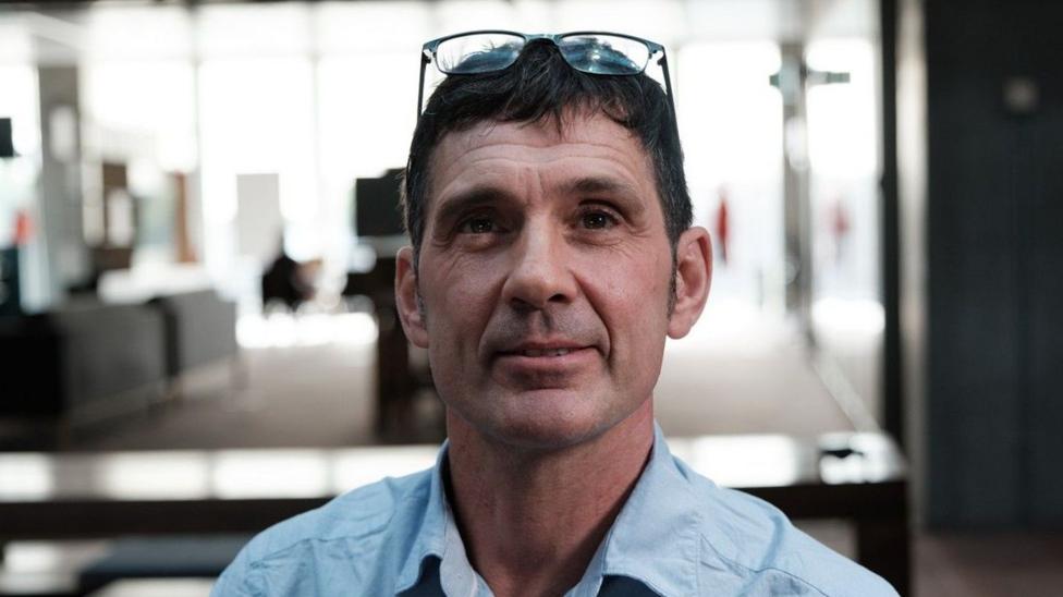 A middle-aged white man looking at the camera. He is wearing glasses on his head and a blue shirt. He has black hair.