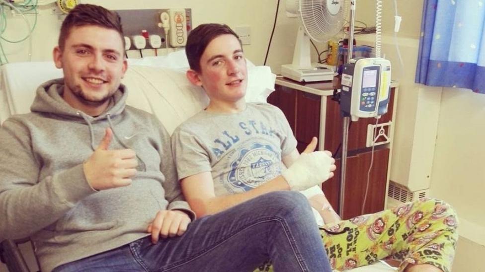 Two people are sat on a hospital bed with their thumbs up. The teenager on the right has a bandage on his hand and has a medical machine next to him. He is wearing a grey t-shirt and smiling. The young man on the left is wearing a grey hoodie and jeans and smiling.