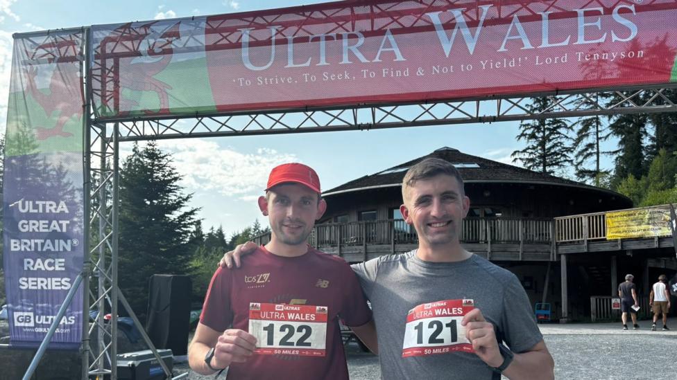 Drew and Saul have their arms around each other and are standing in front of the Ultra Wales race start line. They are holding up their race numbers. The man on the left is wearing a red t-shirt and red cap and the man on the right is wearing a grey t-shirt.