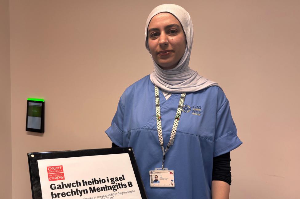 Dalia in a blue medical top is looking at the camera. She is wearing a grey headscarf and is standing in front of a sign advertising Meningitis B vaccination drop-in sessions at Cardiff University.