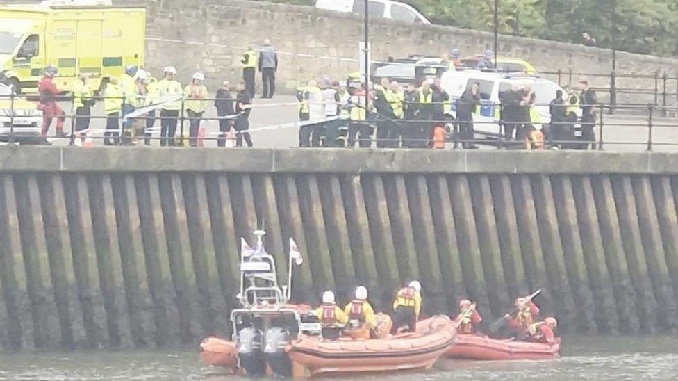 More than a dozen people, most wearing high-viz jackets are along the river bank in Sunderland, where a metal railing is missing and is covered in police tape There are emergency vehicles parked nearby and RNLI volunteers on two orange rescue boats in the river.
