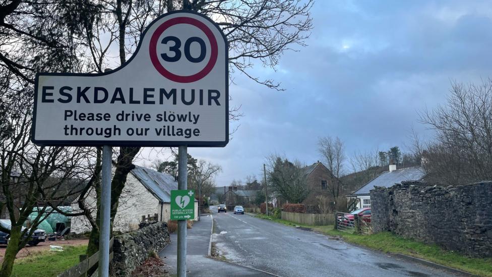 The entrance to the village of Eskdalemuir with a road sign asking people to drive carefully and a 30mph limit. There are houses on both sides of the road and a couple of cars in the distance under grey sky.