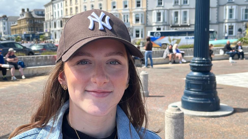 Tilly, a woman wearing a brown baseball cap and a denim jacket, smiles at the camera. Behind her are the tall seafront buildings of Llandudno and a parked coach