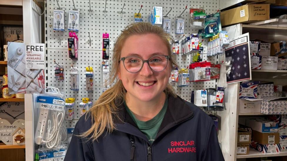 Joanne, a woman with blonde hair wearing a navy blue jacket, smiles as she stands in a hardware store. Behind her are various lightbulbs and extension cables