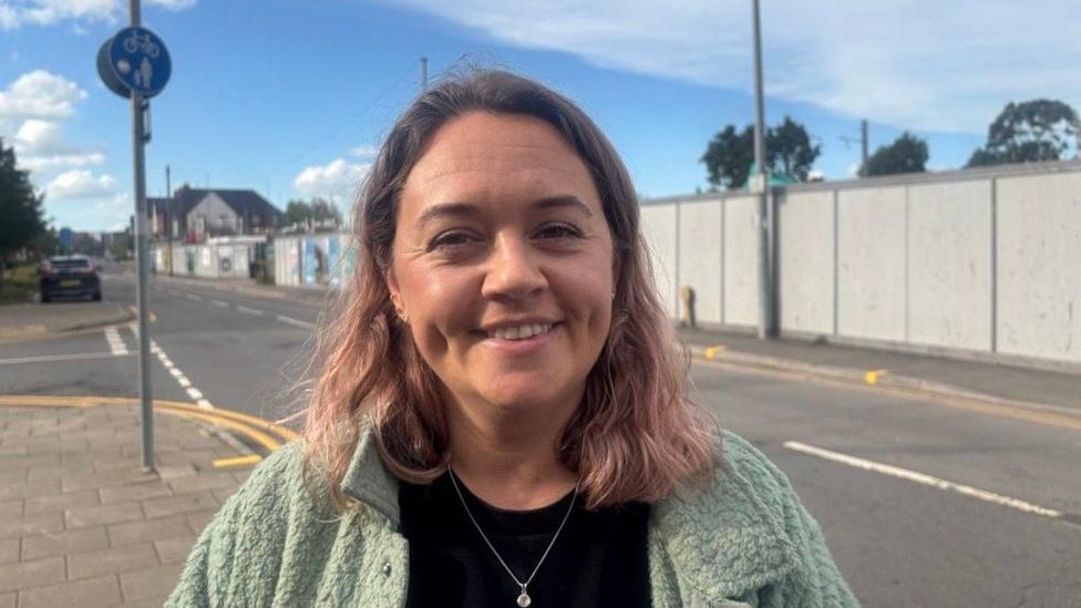 Rhiannon Simmonds stands on the street. She has shoulder length brown hair, wears a black t-shirt and a sage green jacket. Blue sky can be seen behind her.