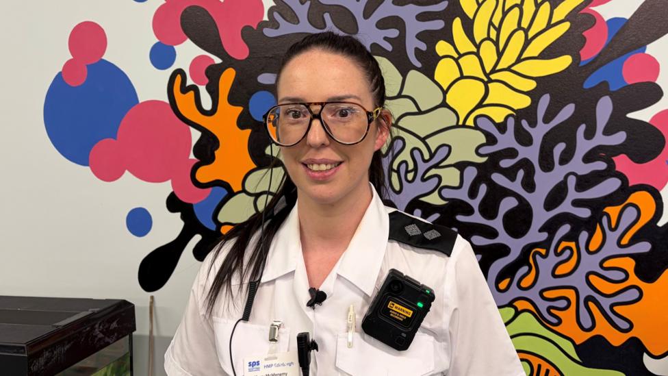 Kerry McMenemy is smiling at the camera. She has long, dark hair in a ponytail and is wearing a white prison uniform. She has an identification badge and a radio attached to her shirt. She is standing in front of a bright, multi-coloured mural that looks like some kind of plant or coral