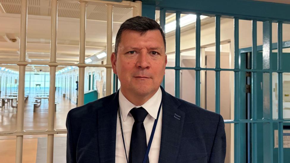 Scott Watson is standing in front of prison bars with a prison corridor and table behind. He has short, dark hair and is looking at the camera. He is wearing a dark suit and tie with a white shirt and a lanyard.