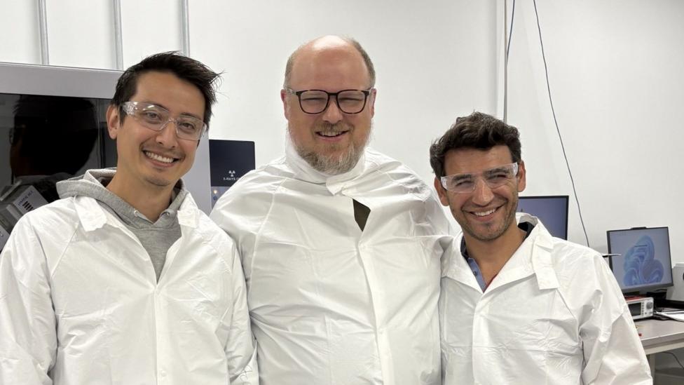Smiling, the three founders of Emerald Battery Labs are standing next to each other wearing white lab coats.