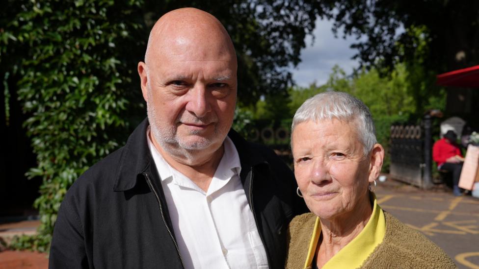 Mark and Roberta Harrison stand in front of a park in Norwich. Mark is wearing a black jacket and a white shirt. Roberta has short, cropped grey hair and hooped earrings, she is wearing a yellow top with a mustard-coloured fleece. There is a large hedgerow behind them and a black gate.