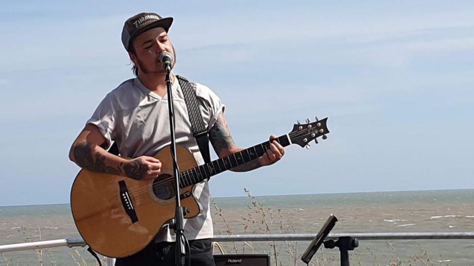 Hobie Harrison performs a gig in Gorleston in Norfolk on the promenade. He is holding a light wood coloured acoustic guitar, which is strapped around his shoulder and he is standing in front of a microphone. He has a baseball cap on and is wearing a grey, v-neck T-shirt. You can see the sea in the background