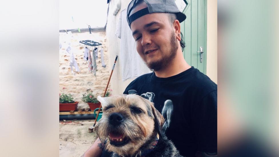 Hobie Harrison sits in front of his parents yard with plants in potted containers behind him. He is wearing a baseball cap the wrong way around and has a black T-shirt on. He is looking at a little terrier type dog which has tan and black colouration