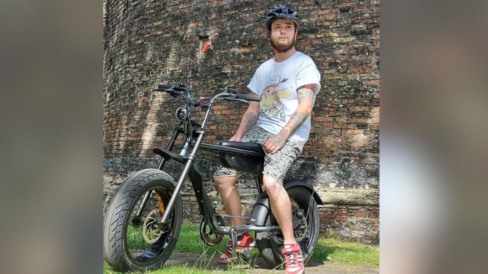 Hobie Harrison sits on his bike. It has huge tyres and looks as if the frame has been hand made. It has a metal and black chassis. Hobie is wearing a Pokemon T-shirt and shorts. He has red and black trainers on. He is sitting in front of a wall posing and is looking to the distance. The bike is resting on a path which is next to grass. He is wearing a black helmet