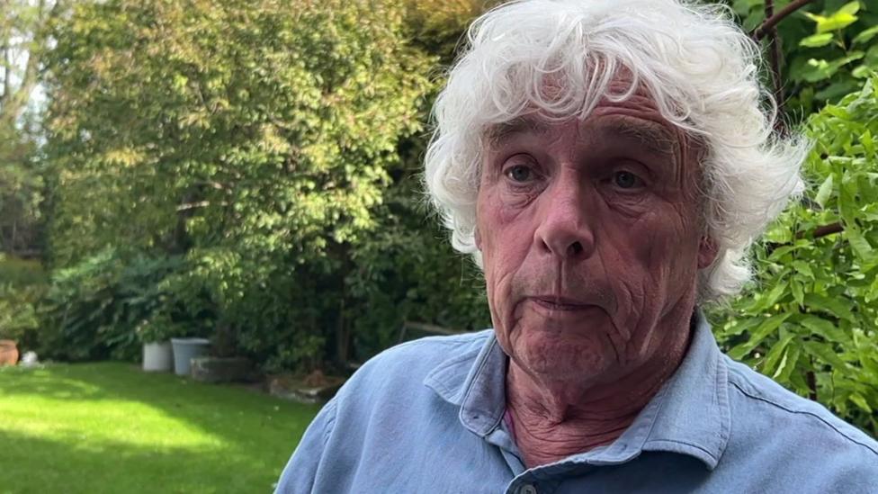 Andrew Illingworth stands on his lawn giving a TV interview. He has curly, white hair and a blue shirt.