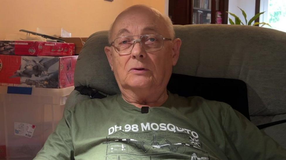 Chris Wheeler sits on his sofa giving a TV interview. He has short, white hair, a T-shirt and glasses.