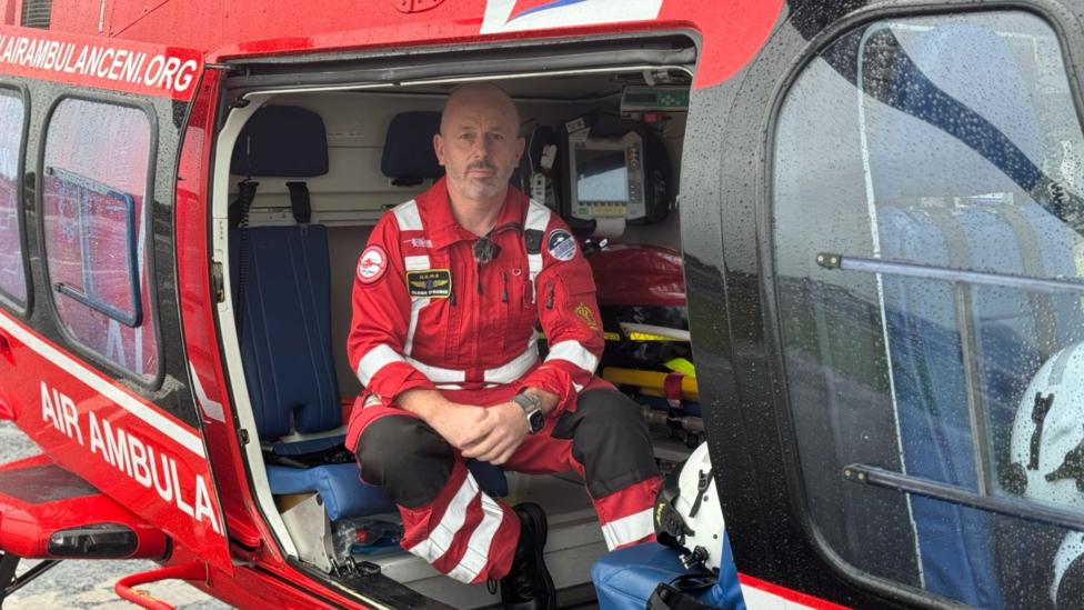 A man is sitting inside the back of a red helicopter looking at the camera. He is bald and has dark and white stubble. He is wearing a red jumpsuit with white stripes. The back of the helicopter contains medical instruments and a stretcher. The helicopter says Air Ambulance.