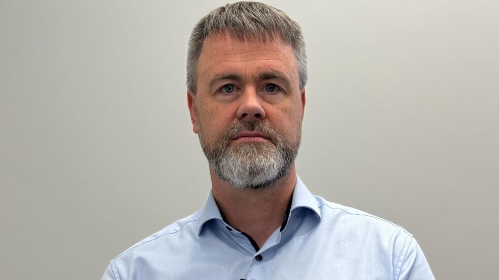 A man looks into the camera. He has short fair hair and a white beard. He is wearing a light blue shirt and is standing in front of a white background.