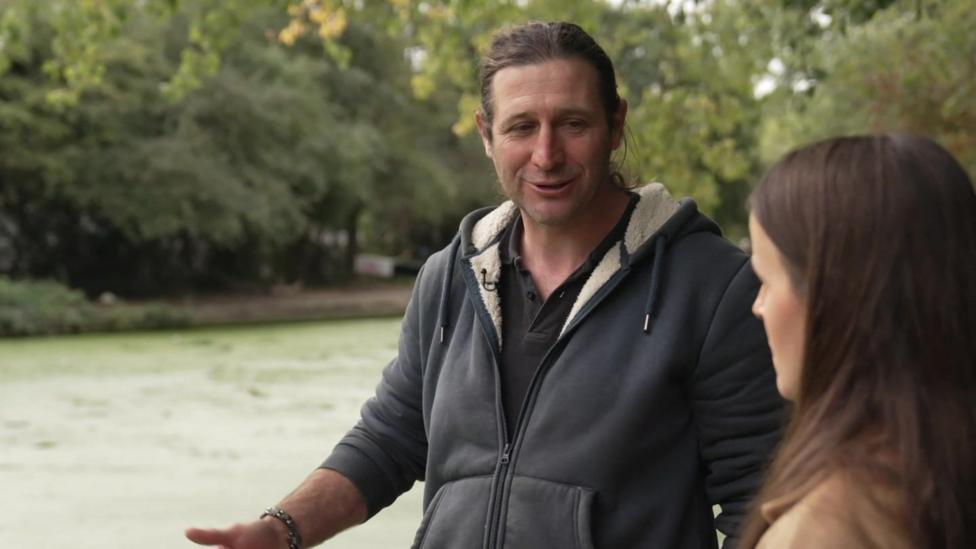 Paul Keane, in a grey hooded fleece, talks to a BBC reporter beside a canal covered in green algae