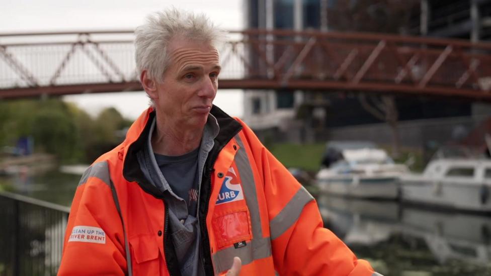 Ben Morris in an orange CURB hi-vis jacket beside the canal, with a red footbridge and moored boats behind him
