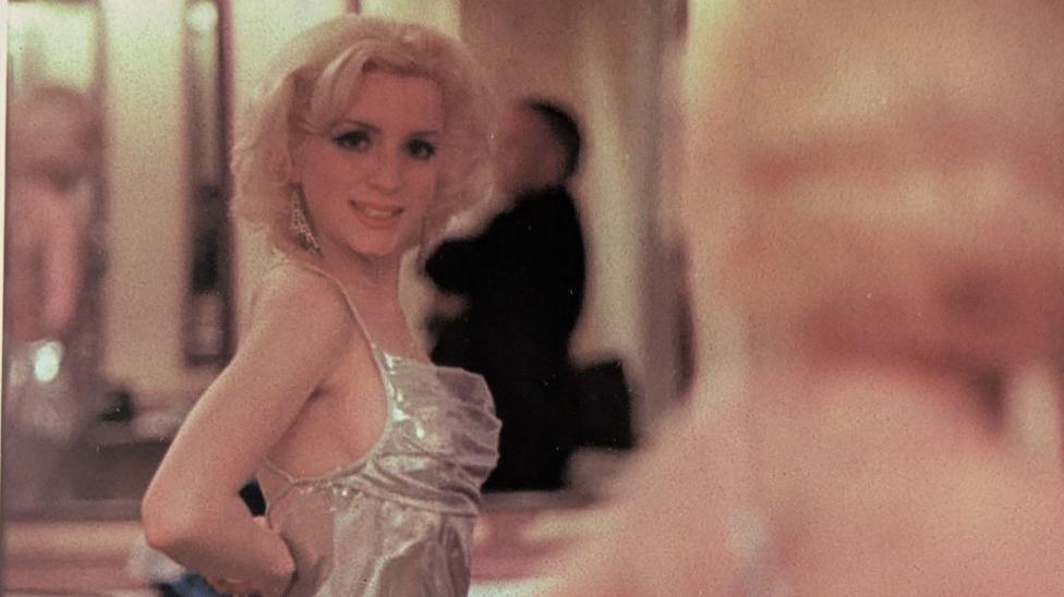 Glamorous young woman looks at herself in the mirror. She has short blond curly hair, a shiny silver dress and long diamond style earrings.