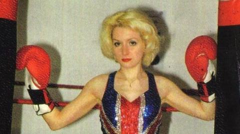 Young blond woman with a stern expression, she has curly short hair and wears a glittery union jack dress. She has red boxing gloves with her hands raised to her shoulders.