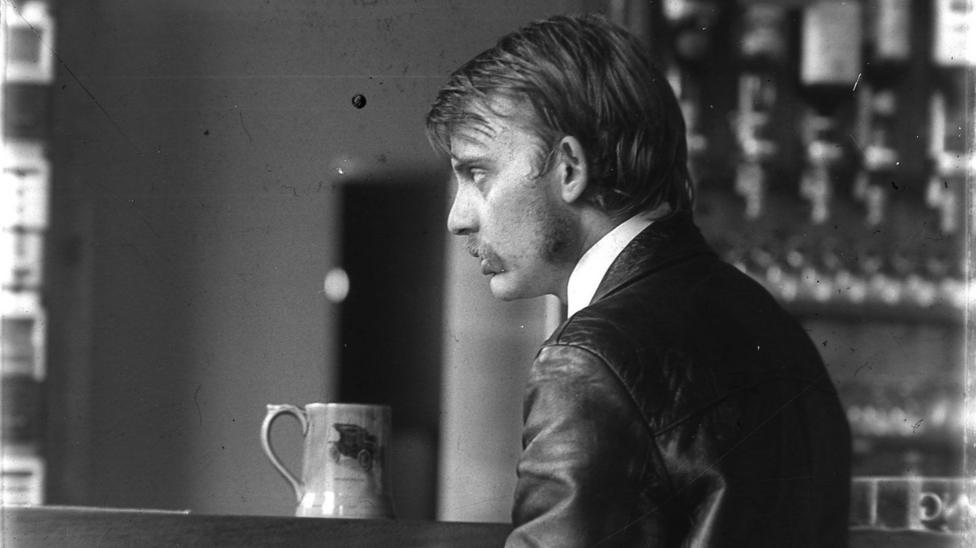 A black and white photograph of Glyn Jones which was believed to have been taken in The Prince Albert pub in Ely, Cambridgeshire, in 1976. He is sitting at the bar with a pint in front of his and is looking towards the bar with his back facing the camera.