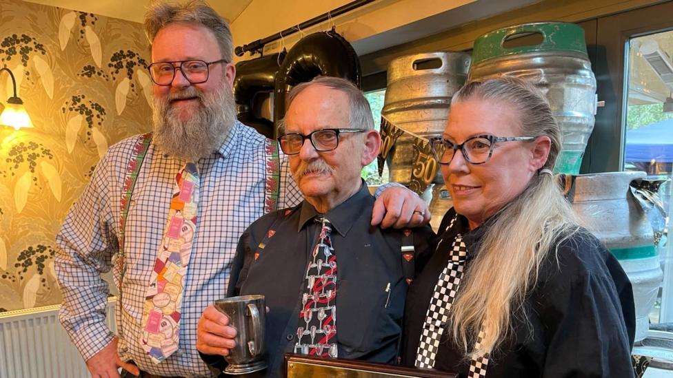 A picture of Mike Connelly, Glyn Jones and Jeanene Connelly standing in a row. They are presenting Glyn with a golden Greene King plaque during a surprise party for Glyn's years of service. Behind them are kegs. All three of them are dressed in a shirt, patterned tie and patterned braces.