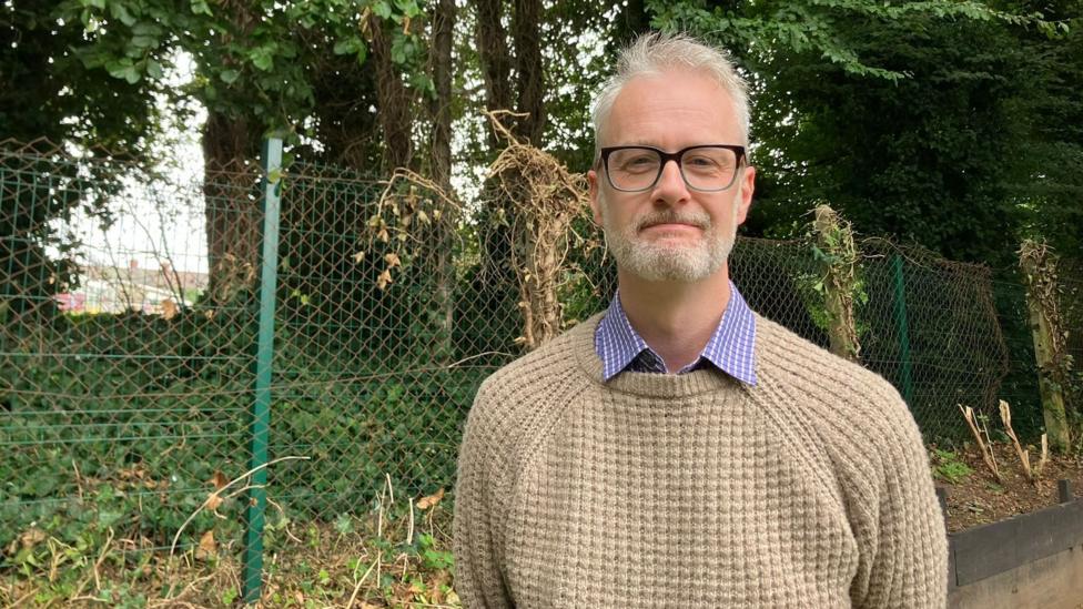 Andrew Ibbotson has grey hair and a grey beard and is smiling slightly to camera. He's wearing glasses and a beige jumper with a blue checked shirt and collar. He is standing in front of a green metal fence with trees the other side of the fence and some branches overhead.