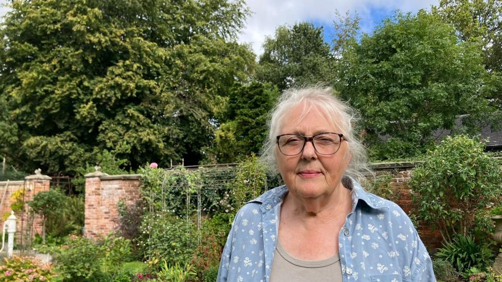 A woman with grey hair tied back and who is wearing glasses. She's wearing a pale blue shirt with a white pattern on it and is standing in her garden, with tall trees in the background