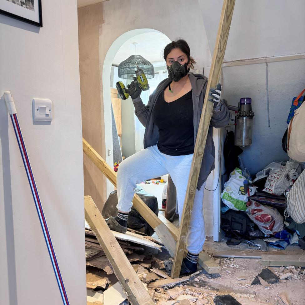Jasmine Gurney wears a protective mask and holds a drill as she stands surrounded by debris in a house.