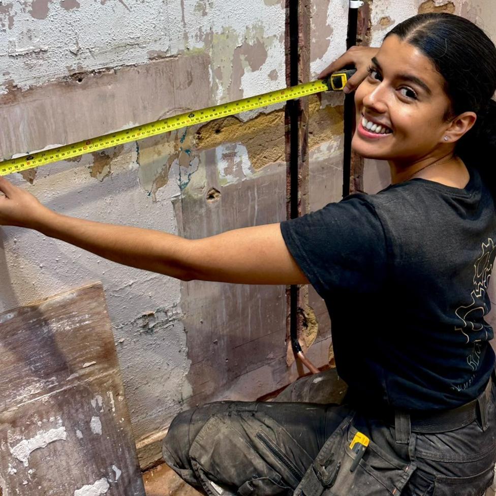 Bliss Cunneen holds a tape measure up to a wall and smiles at the camera.