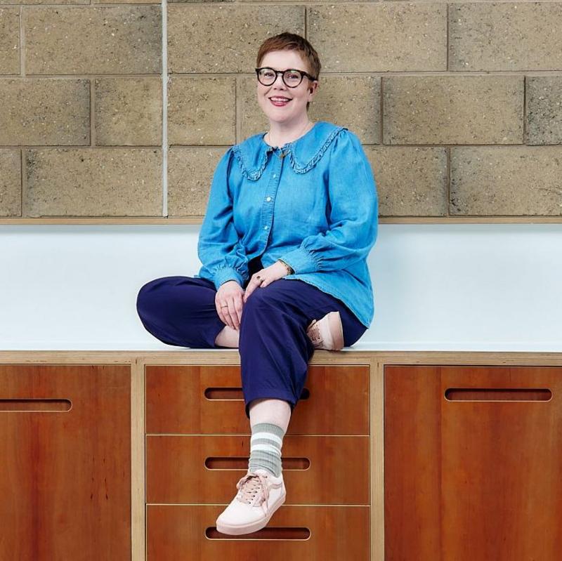 Anna Moynihan, wearing a blue shirt, blue trousers, stripey socks and trainers, sits on a kitchen worktop. She is smiling at the camera and is wearing glasses.