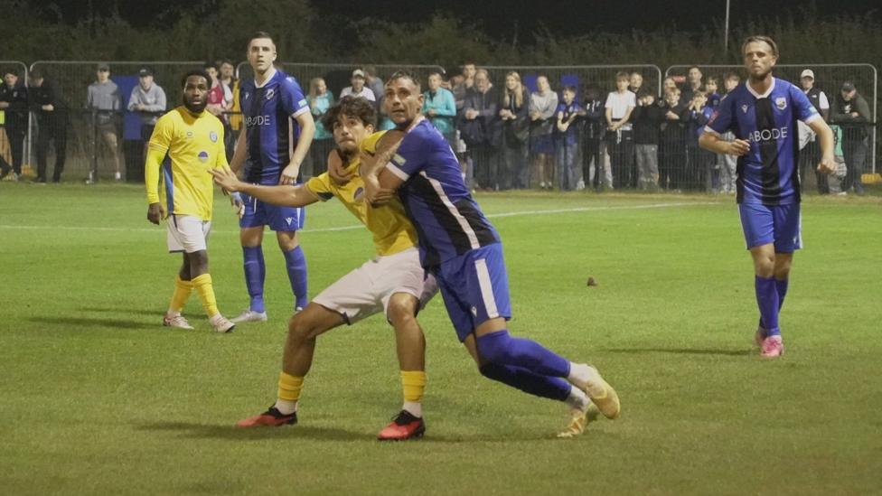 A football player in yellow kit fights off an opponent in blue as team mates watch on