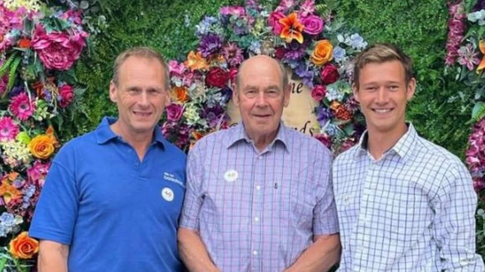 This is a photo of three generations. Peter Philpot (middle), Chris Philpot (left), Thomas Philpot (right) The men on the right and in middle are wearing chequered shirts, the man on the left is wearing a polo shirt. They are standing in front of a flower display.