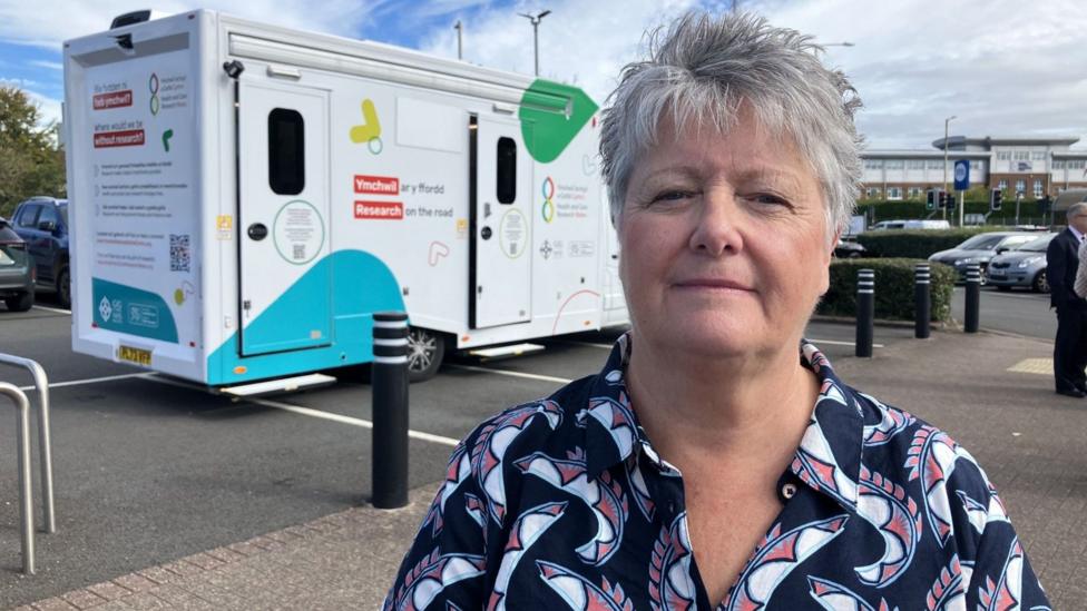 A woman with short hair and a colourful blouse stands in front of a vehicle which is conducting the tests.