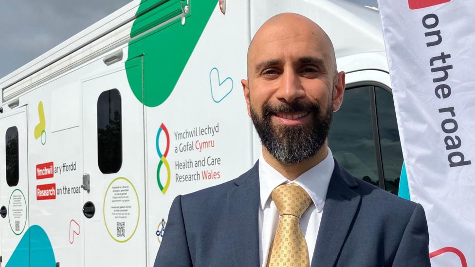 A man with a beard stands in a suit and tie in front of a Health and Care Research Wales vehicle.