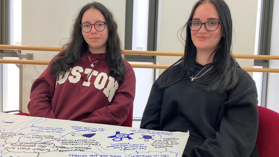 Two teenage girls sit at a table with a large sheet of paper in front of them with blue writing. Both girls are wearing jumpers and have dark hair.