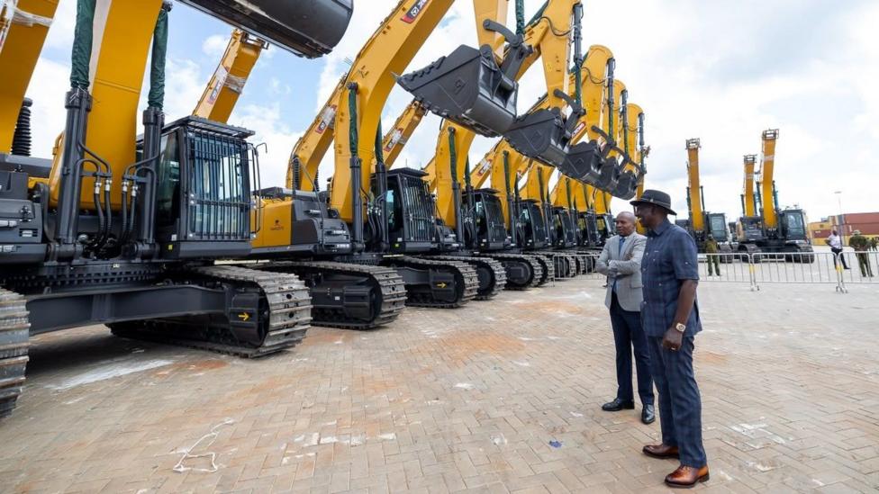 A line of heavy construction excavators parked next to each other on a dirt ground surface. In the foreground is two men in official attire looking at the equipment