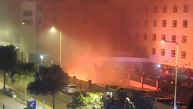A night-time view of a residential street shows a large plume of orange-lit smoke. Streetlights illuminate the scene and the smoke is spreading, partially obscuring buildings and trees. Several cars are parked along the road and in a nearby parking area, while the upper floors and rows of windows of surrounding buildings can be seen through the haze.