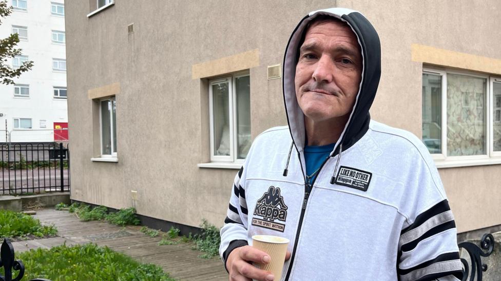 A person wearing a white hooded jacket stands outside a residential building holding a paper cup. Apartment windows, a security sign, railings and a paved area are visible around the building, with part of a larger building visible in the background.