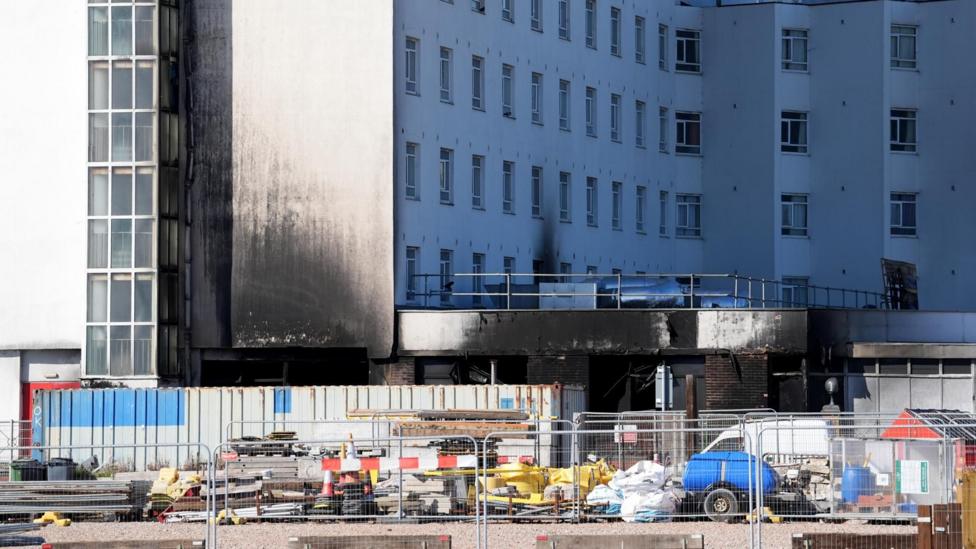 The exterior of a fire-damaged building is heavily blackened by smoke, with dark scorch marks extending up walls and around window openings. Construction equipment and containers are positioned in front of the damaged section.