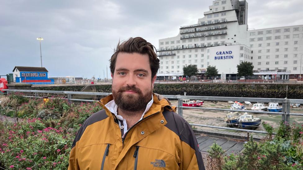A person wearing a brown waterproof jacket stands on Folkestone Harbour Arm. The Grand Burstin Hotel is visible across the harbour in the background, alongside moored boats, railings, flower beds and the Harbour Arm sign under a cloudy sky.