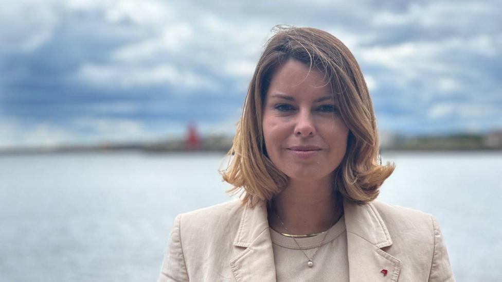 North East mayor Kim McGuinness, in North Shields. She has blonde hair, styled in a bob, and wears a cream top and blazer. She is smiling. The sea lies behind her, with a blurred coastline in the distance.