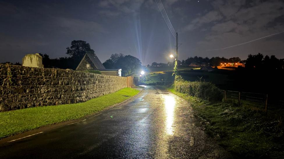 A road lit up with car lights in a rural location. There are puddles on the road with a church and stone wall next to it.