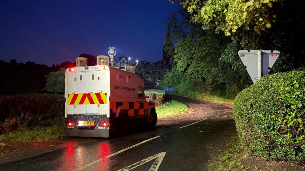 A police Land Rover sits on a rural road, it has yellow and red stripes and a blue an yellow checked pattern.